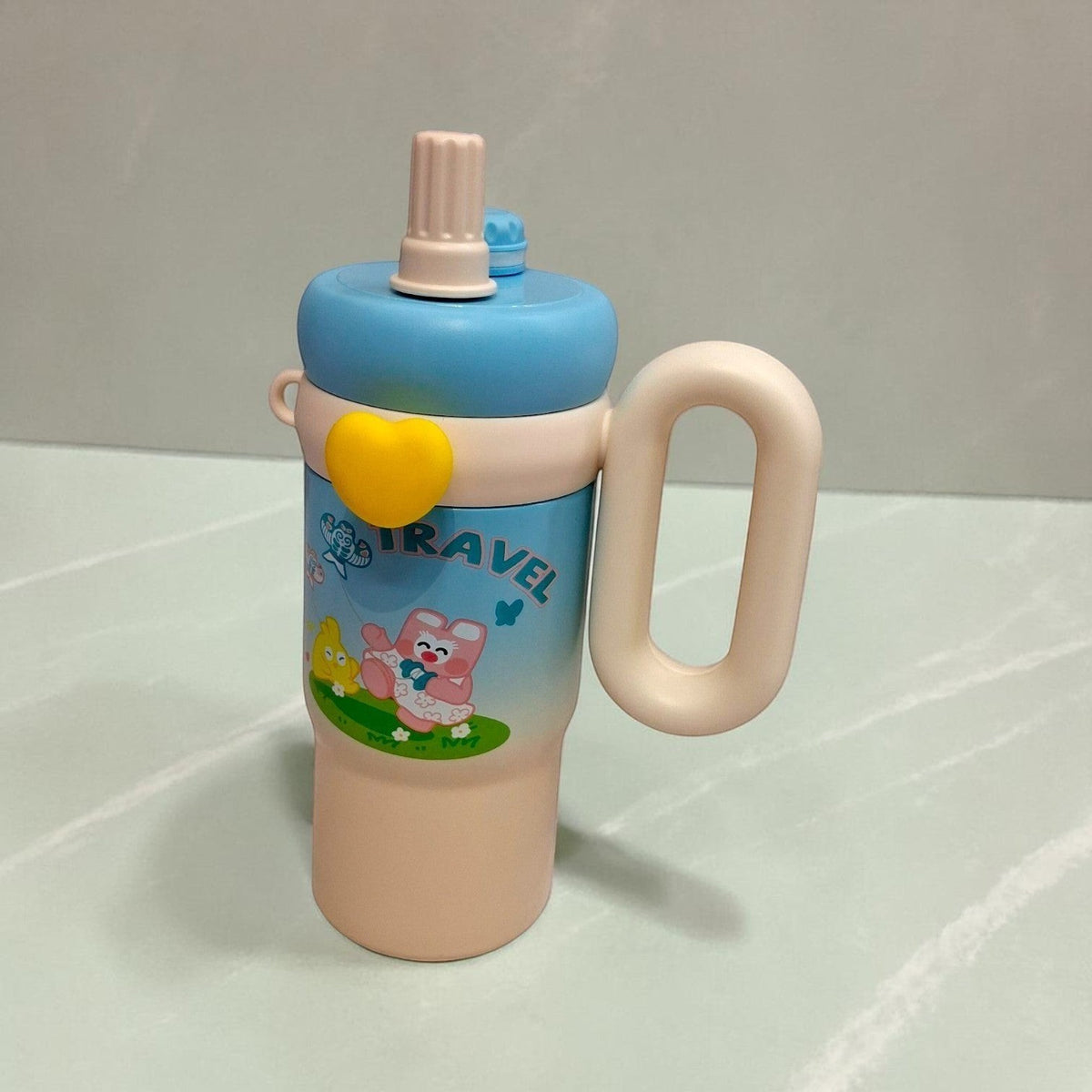 Tiny Tokens pastel blue and cream kids travel tumbler with handle, straw, and yellow heart charm. Features cartoon animals and the word TRAVEL.