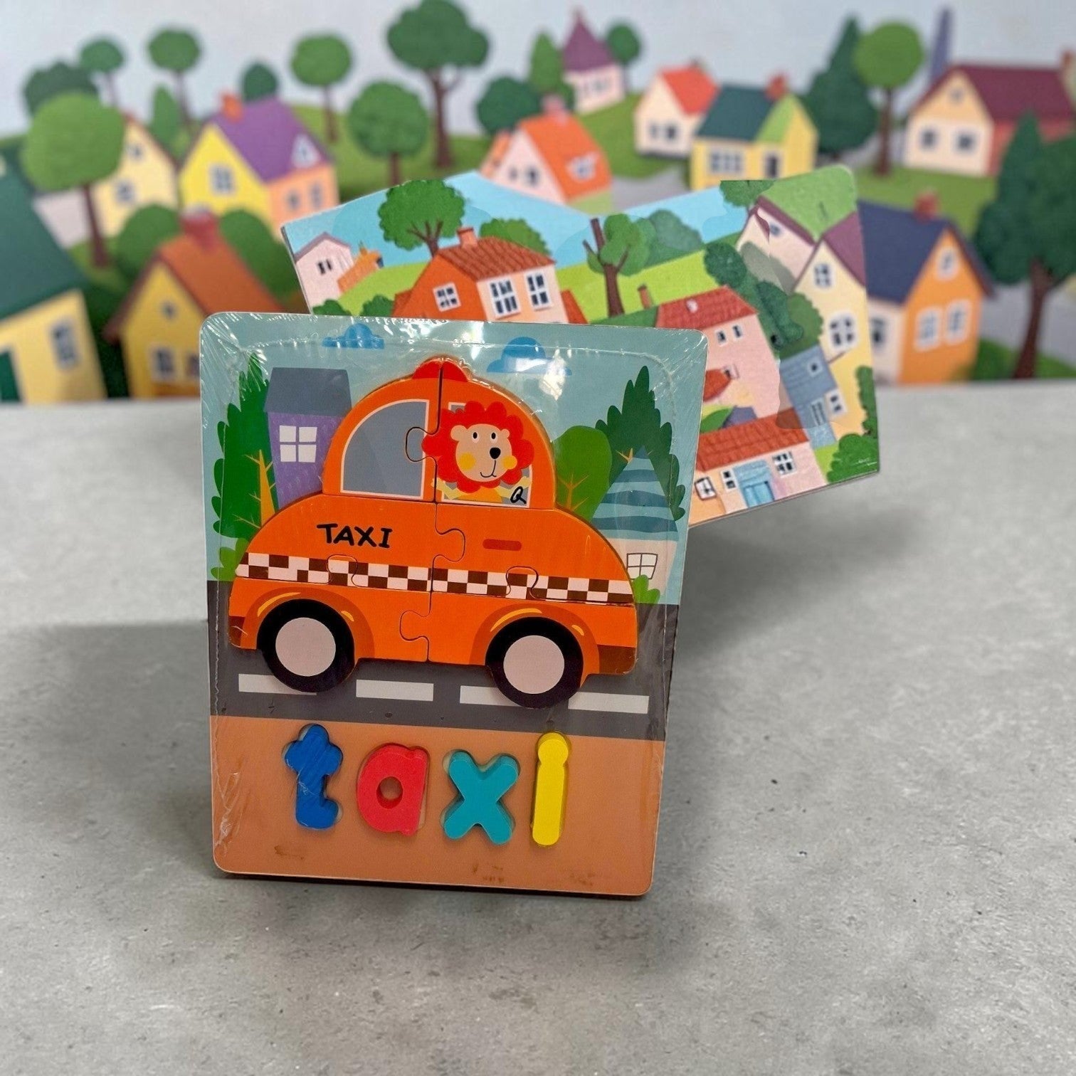 Tiny Tokens orange wooden taxi puzzle board with chunky letter pieces for spelling 'taxi'. Features a lion driver and city background.
