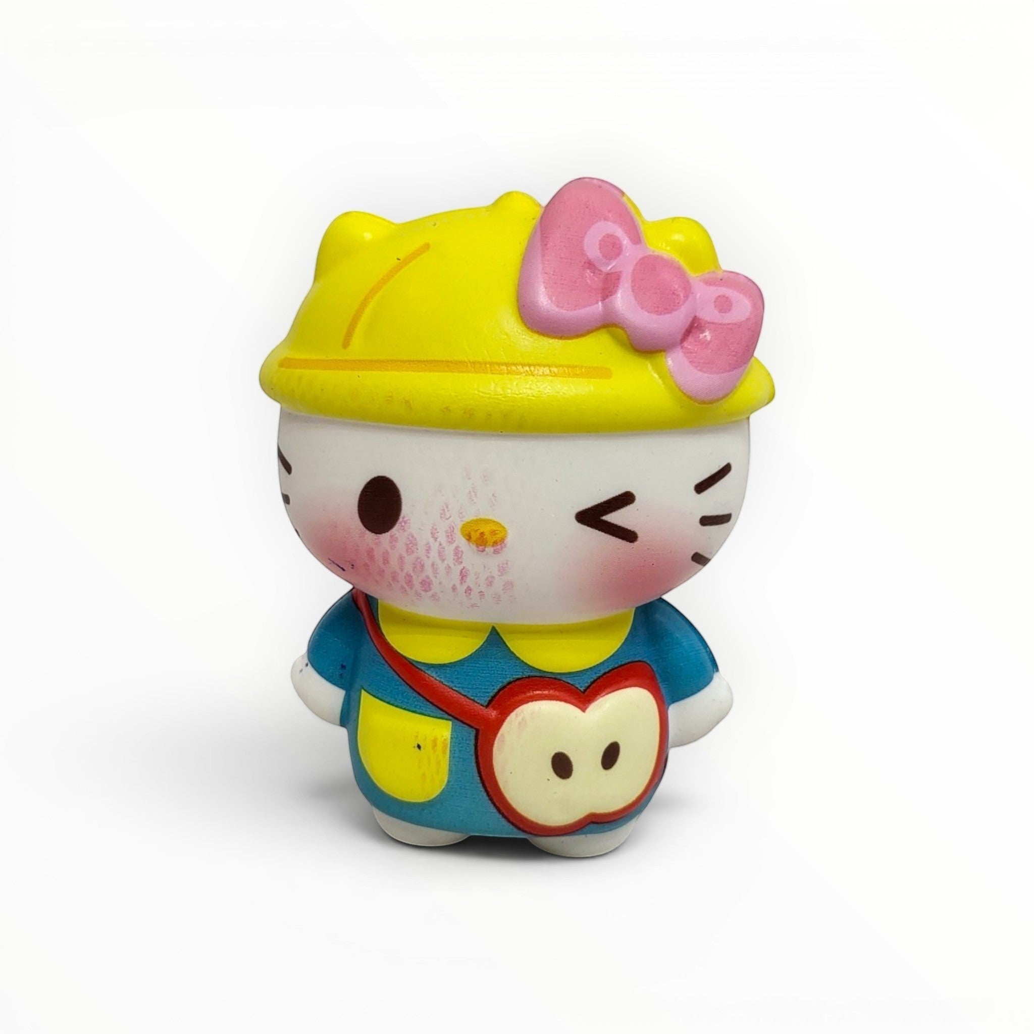 Tiny Tokens white cat squishy toy with yellow hat, pink bow, blue outfit, and apple-shaped satchel. Winking expression.